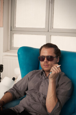 Painter Andreas Golder wearing sunglasses, seated in a turquoise armchair in front of a bright window 