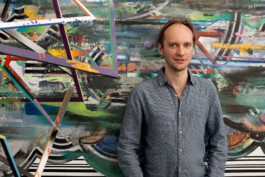 Painter Christian Achenbach in a light blue shirt, standing in front of a large colorful abstract painting with diagonal stripes 